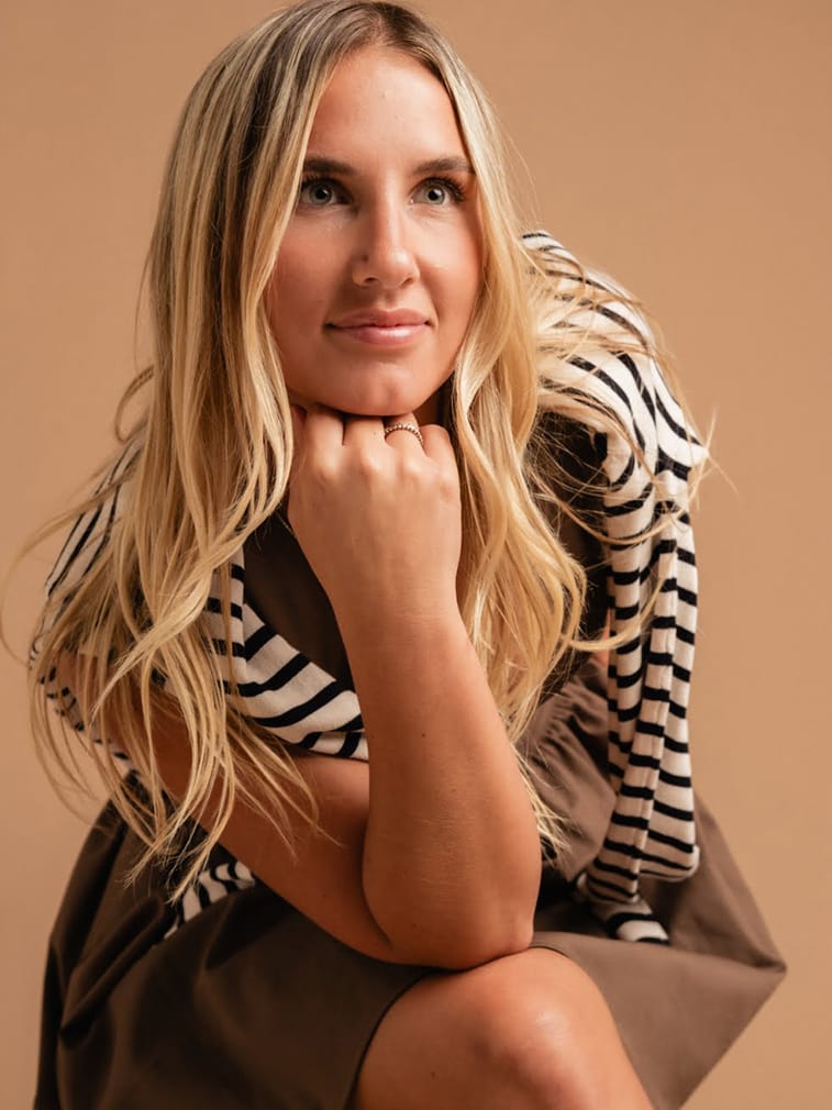 A blonde woman in a striped dress posing with her hand on her chin.