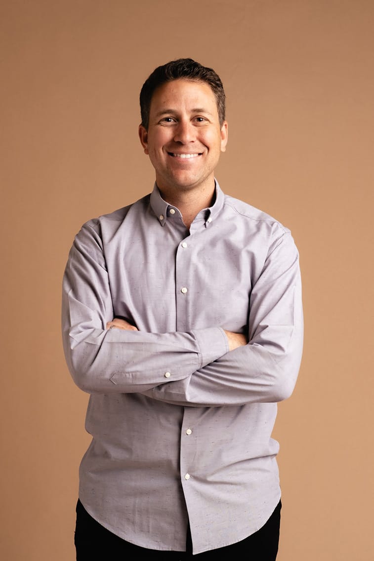 A man in a gray shirt smiling in front of a brown background.