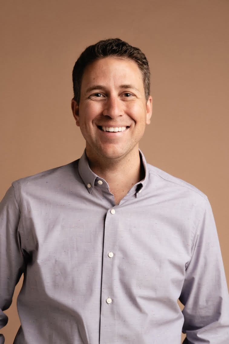 A man in a gray shirt smiling in front of a brown background.